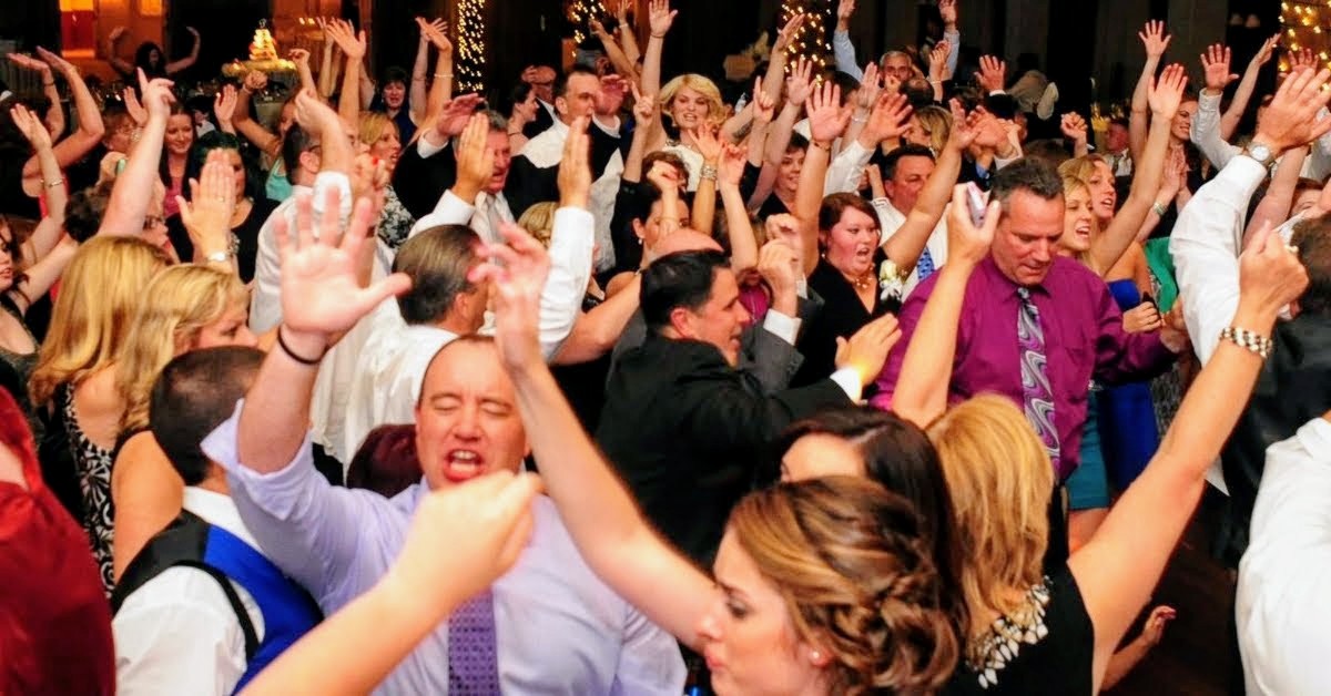 Packed reception dance floor with dozens of guests waving both hands in the air
