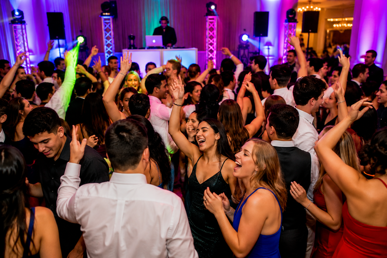 A crowded ballroom dance floor with hands up and purple uplighting