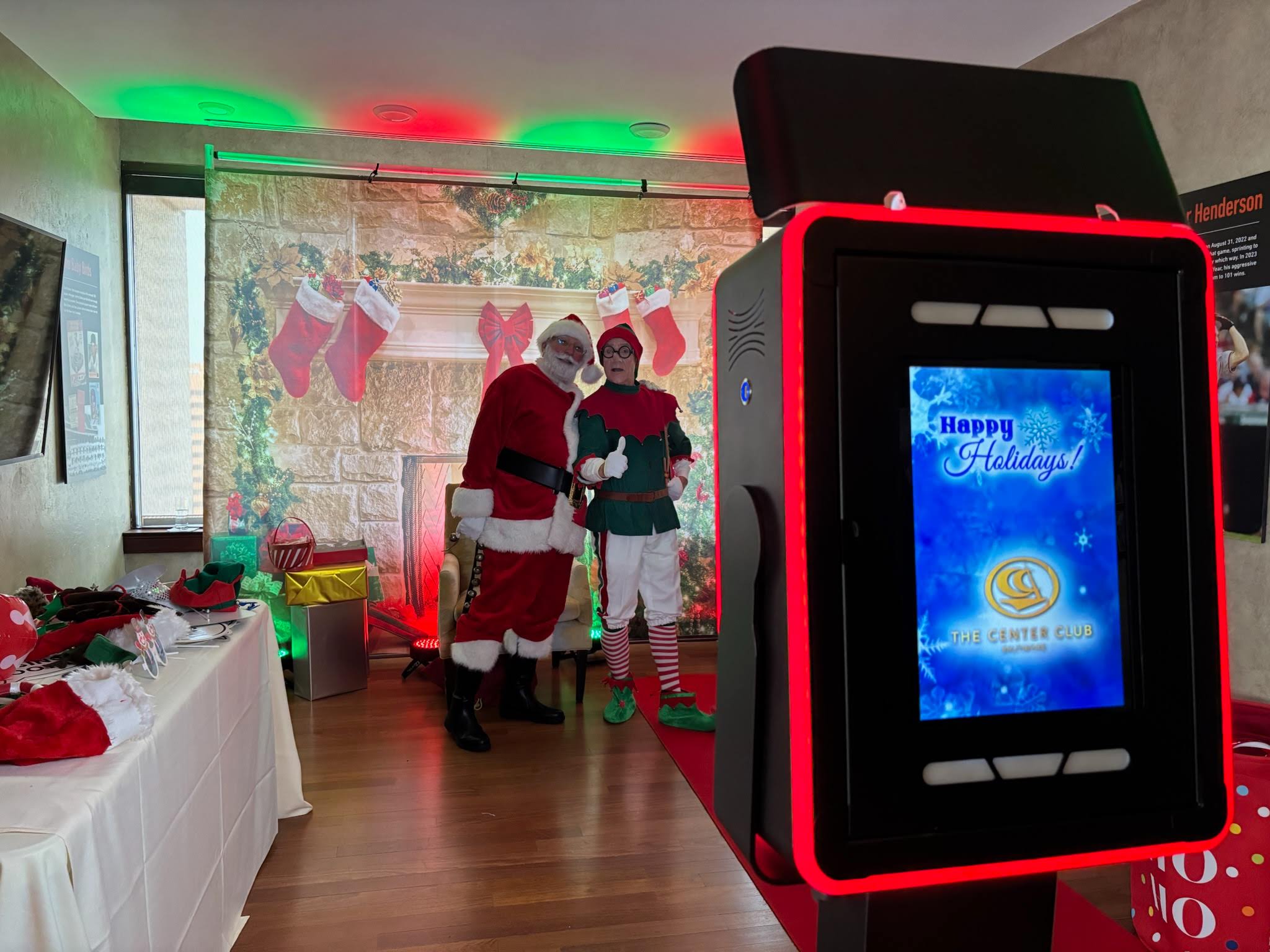 Santa and an elf posing at a holiday photo booth in front of a fireplace backdrop