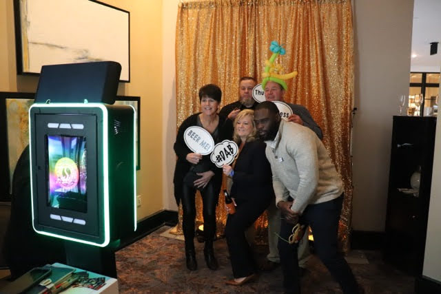 Five guests leaning in together with prop signs at a gold sequin backdrop