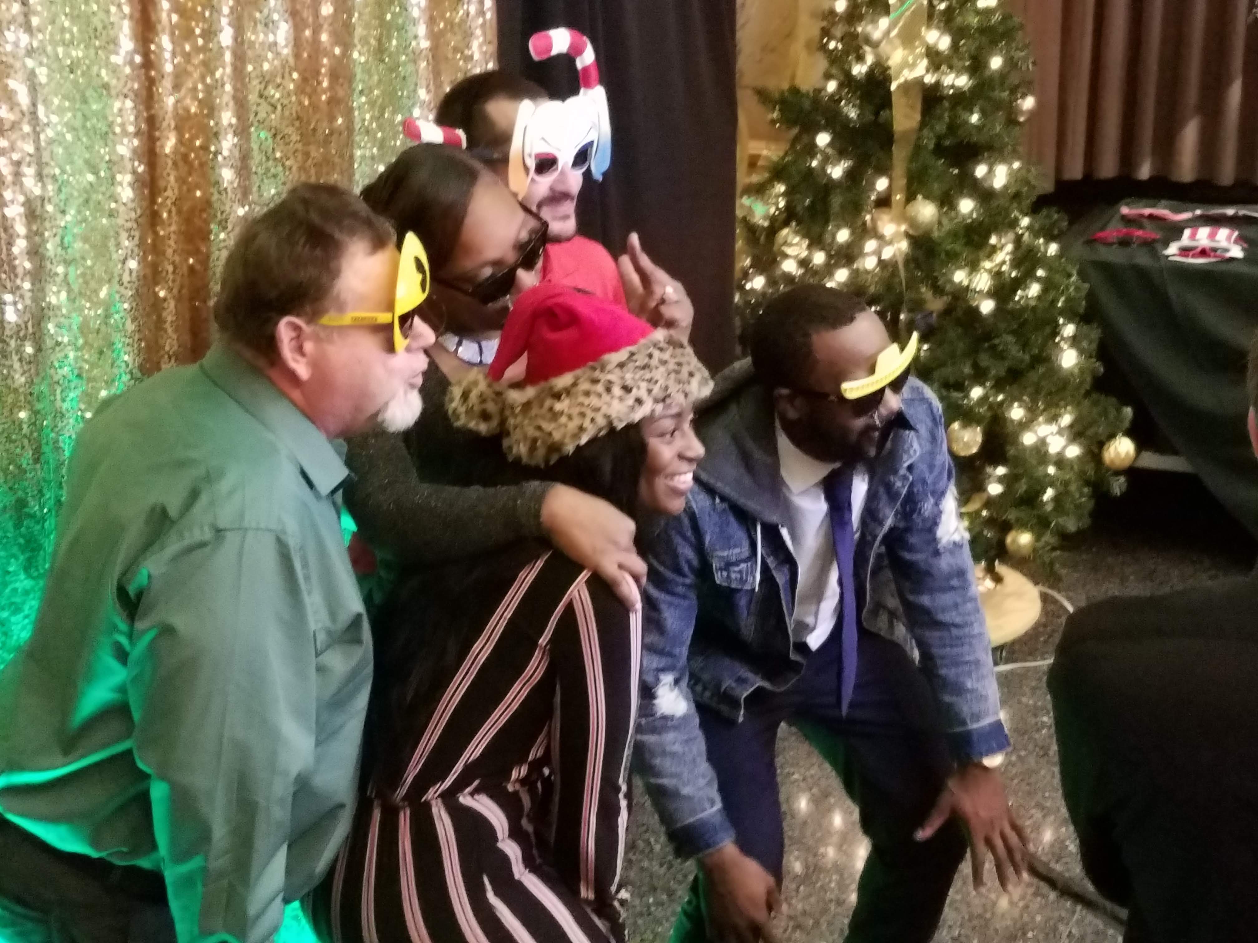Guests in novelty glasses and hats crowding together at the photo booth, gold sequin backdrop and a lit Christmas tree behind them