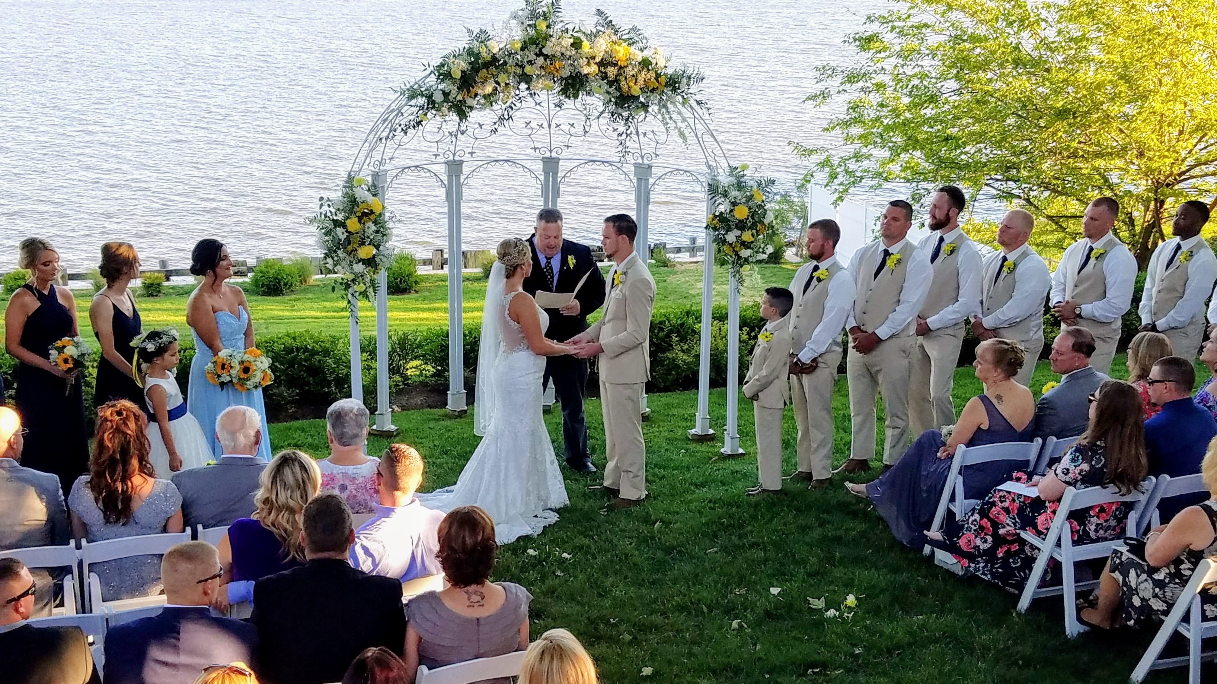 Outdoor waterfront wedding ceremony, the couple holding hands under a floral arch as guests watch