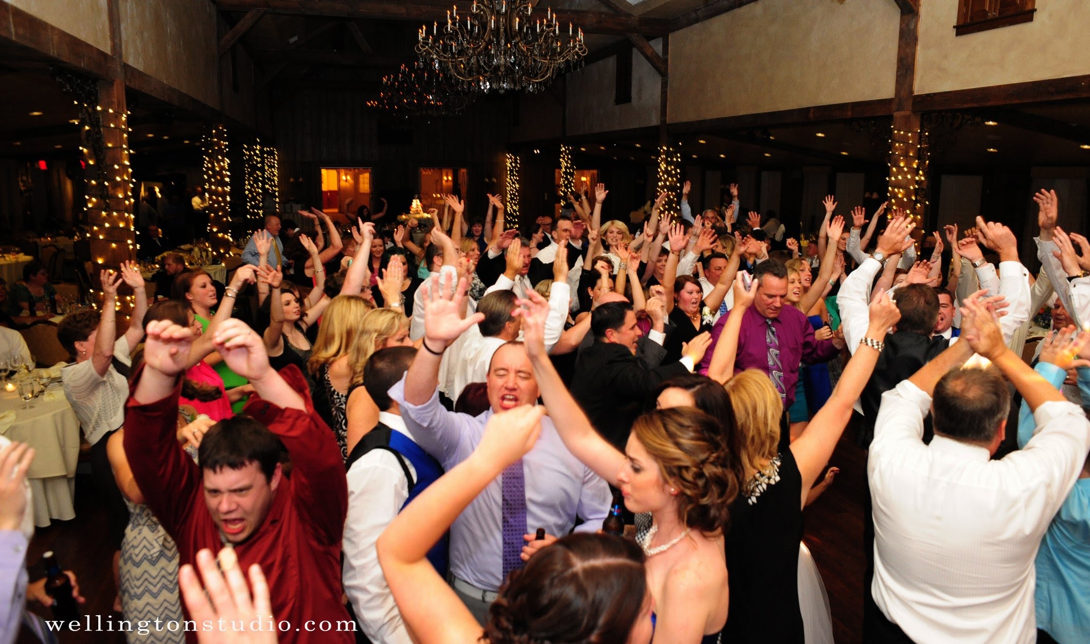 Packed wedding reception dance floor under a chandelier and string lights, guests with both arms raised