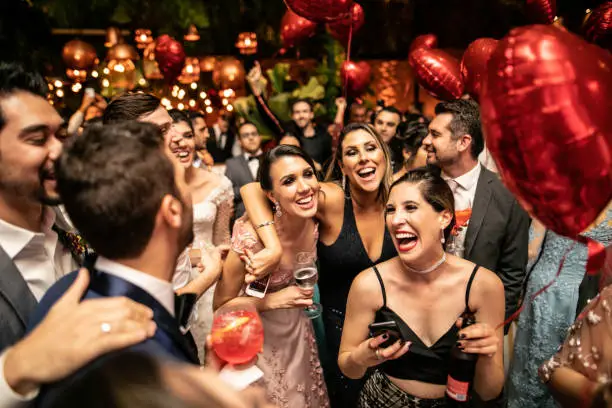 Wedding guests in evening wear laughing together under red heart balloons at an outdoor reception