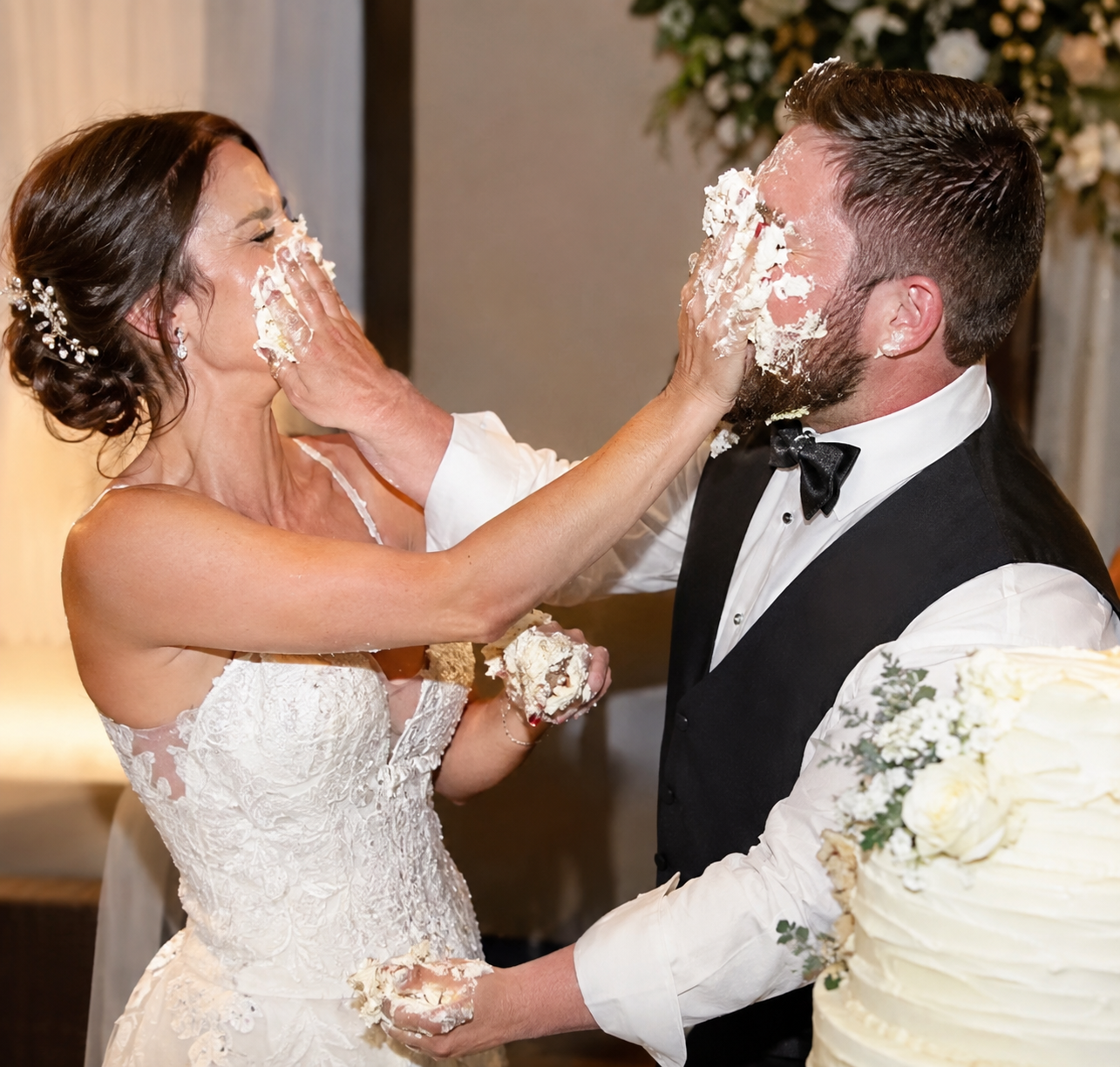 A bride and groom in a white gown and black tuxedo vest smash handfuls of white wedding cake into each other&rsquo;s faces beside the cake