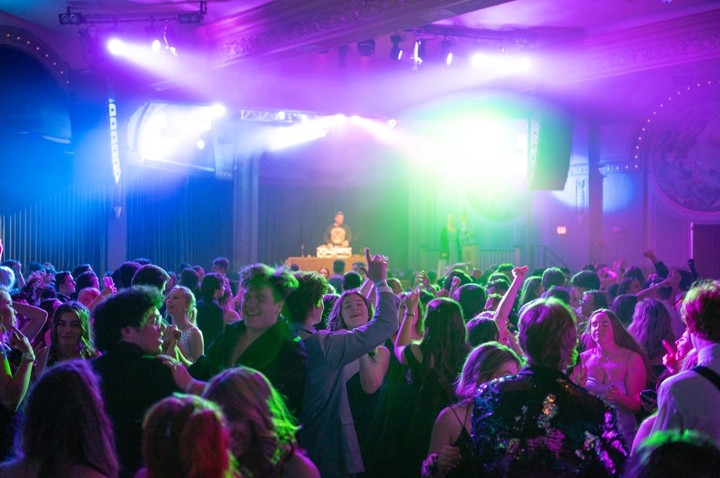 Students in formal wear packing the dance floor with their hands in the air at a prom while the DJ plays on stage behind them